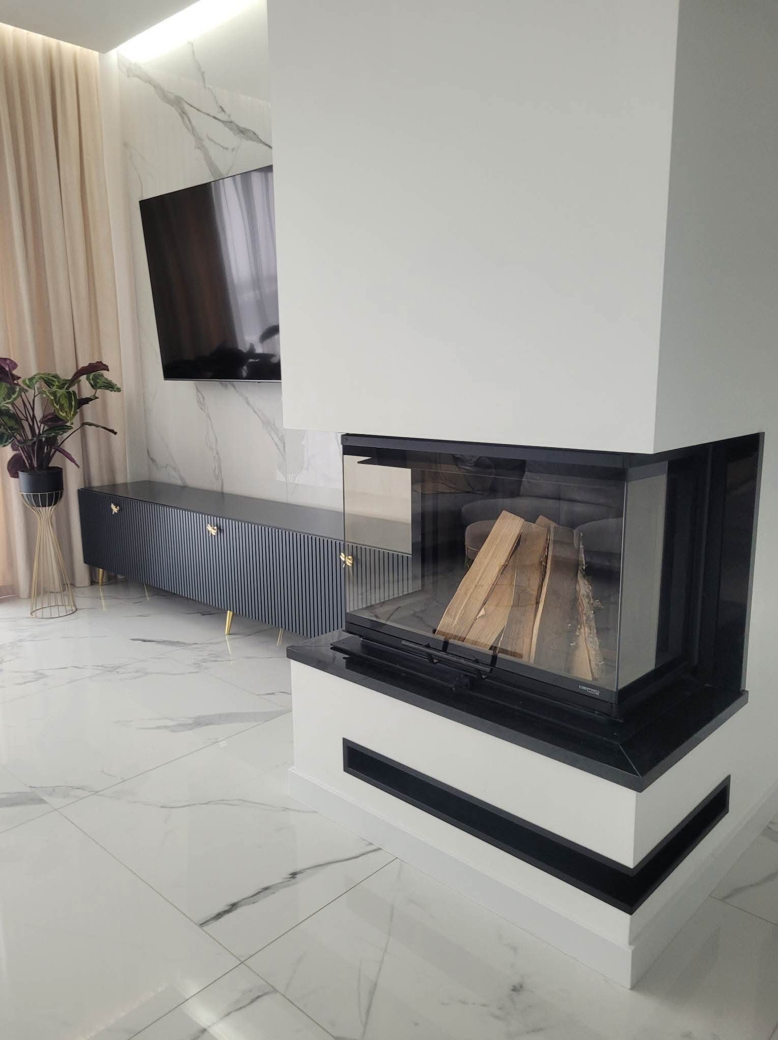 Nowoczesny kominek narożny z szybą, drewnem w środku i czarnymi akcentami, na tle marmurowej ściany z telewizorem i szafką RTV w salonie.