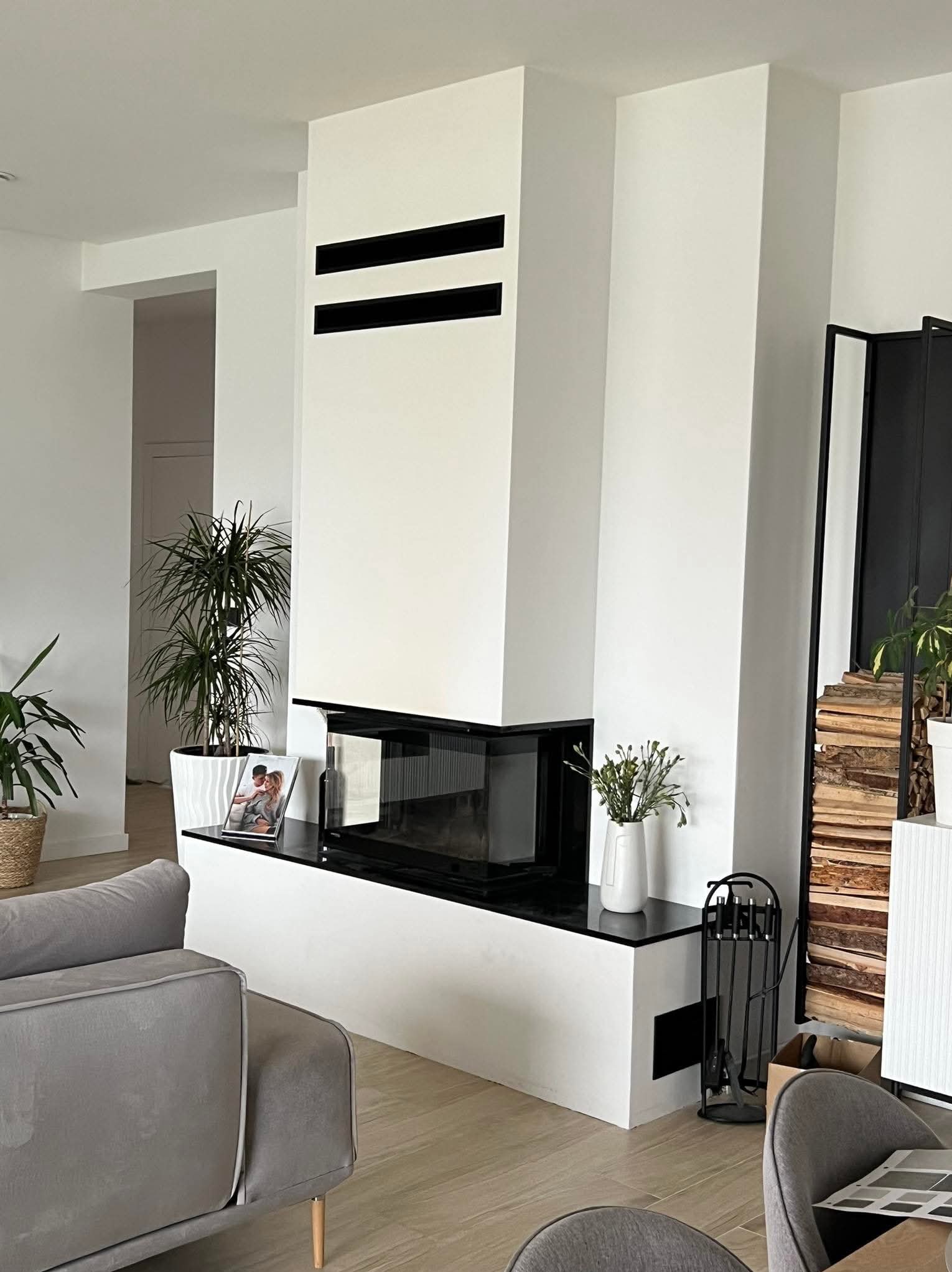 Nowoczesny kominek w salonie z czarnym blatem i elementami dekoracyjnymi: rośliny, zdjęcie w ramce, drewno. Minimalistyczny design, jasne wnętrze.