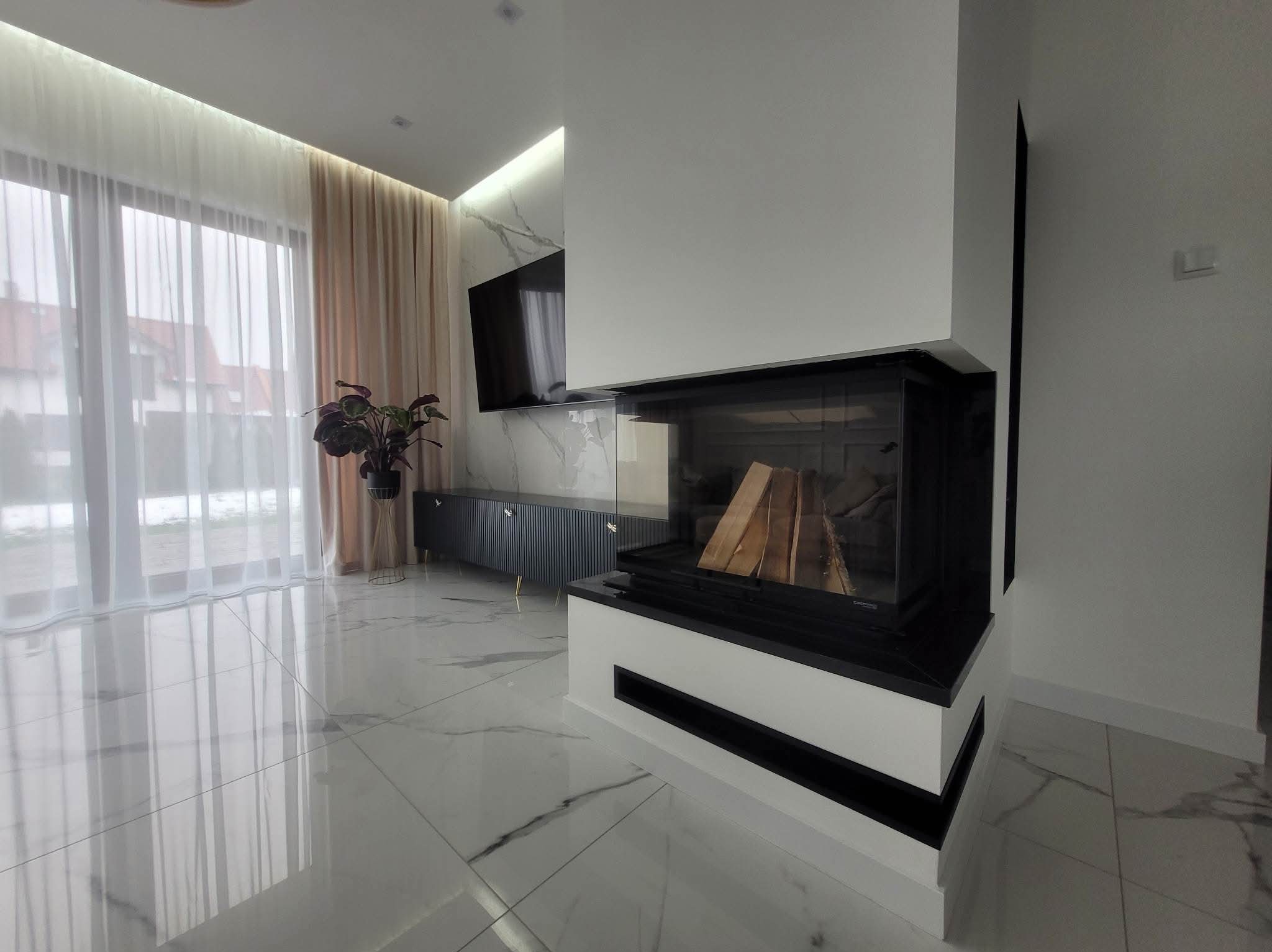 Nowoczesny kominek narożny z szybą, obudowany białym kamieniem, z widocznym drewnem w palenisku. W tle marmurowe ściany i podłoga, minimalistyczny salon z TV.