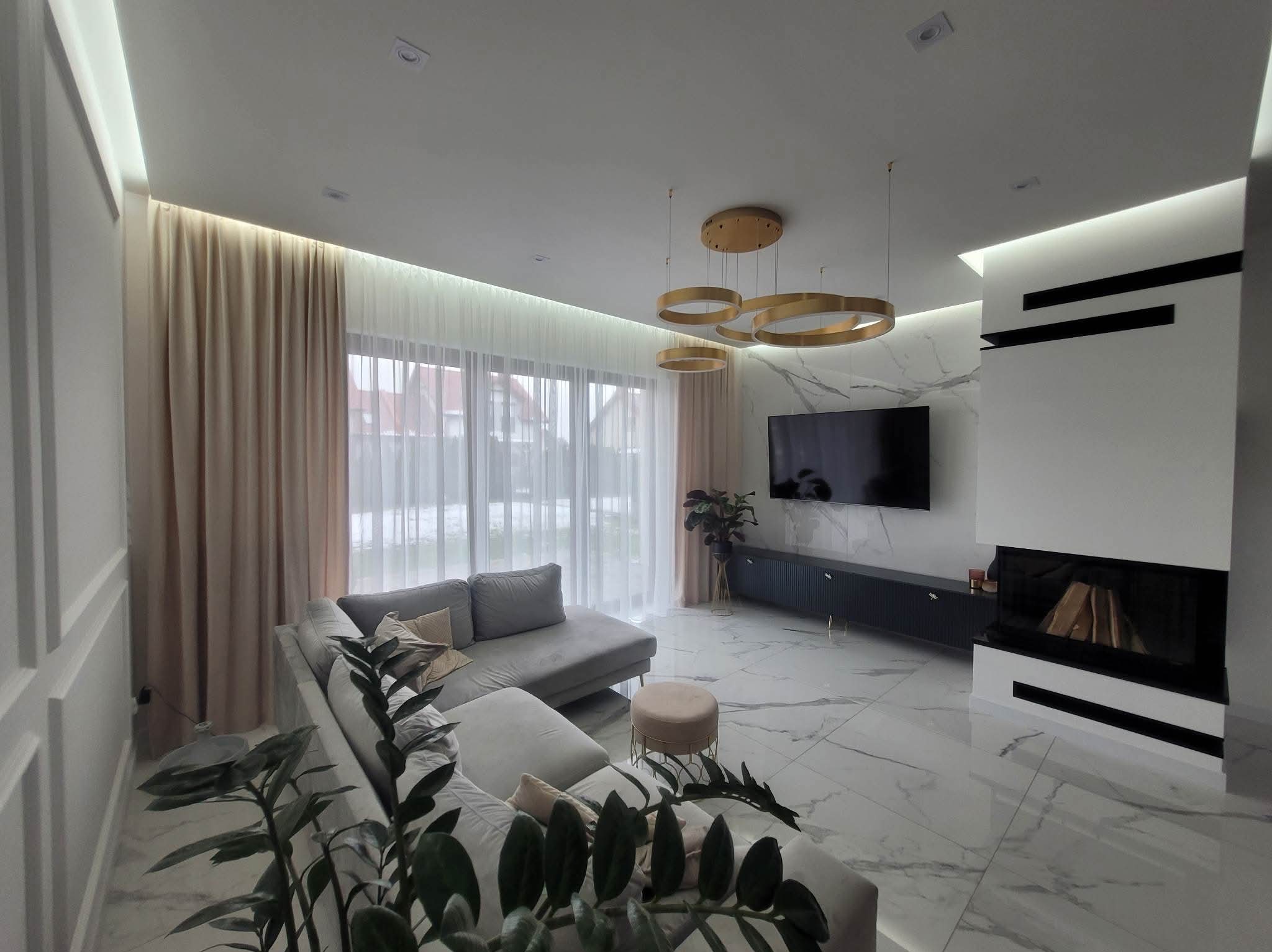 Elegancki salon z kominkiem, marmurową ścianą i podłogą. Nowoczesne oświetlenie, szara sofa i zasłony tworzą spójną całość. Widok na zewnątrz przez okno.