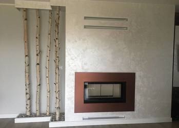 Nowoczesny kominek z obudową w odcieniach brązu, dekorowany brzozowymi gałęziami i srebrzystą ścianą. Minimalistyczny design z akcentami natury.