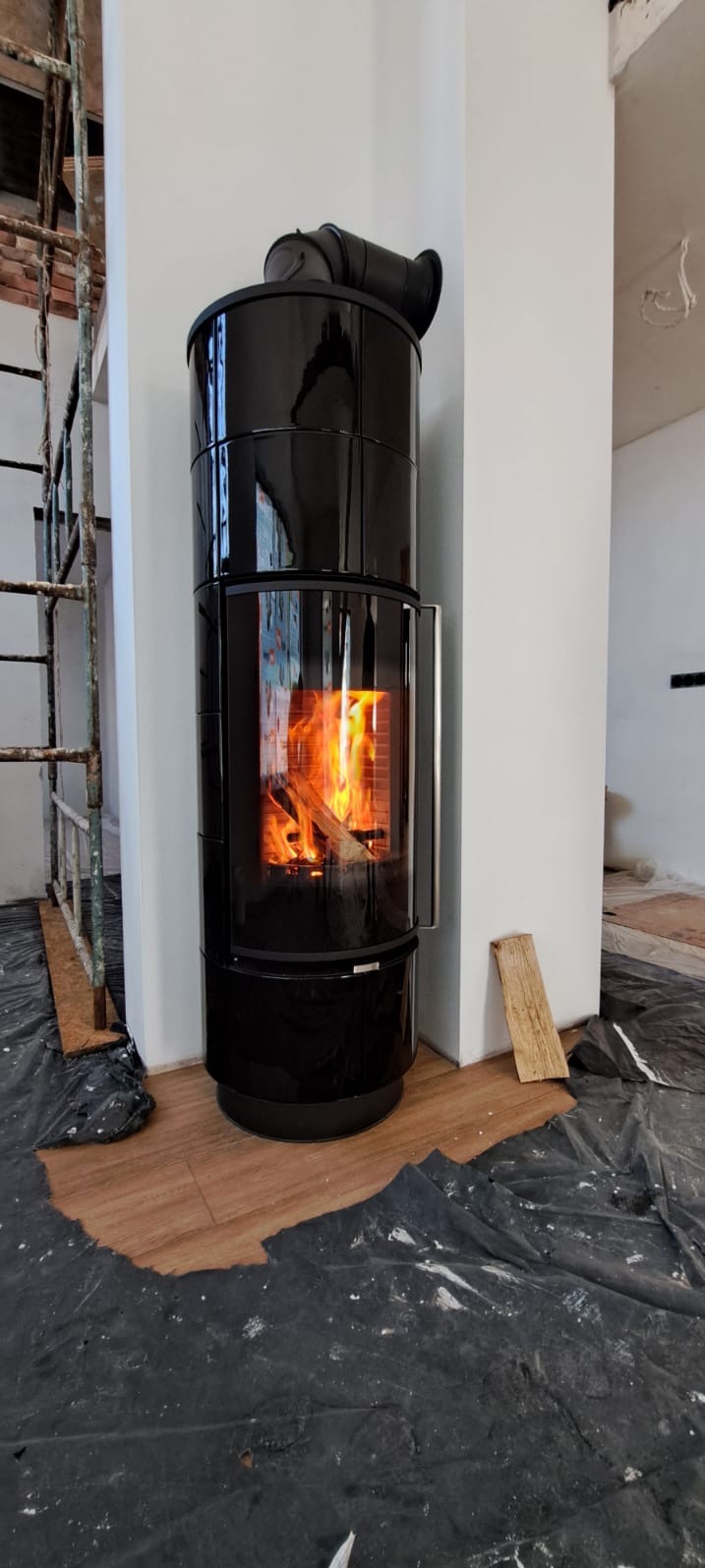 Czarny, cylindryczny kominek wolnostojący z paleniskiem, otoczony folią ochronną i elementami rusztowania, w trakcie prac wykończeniowych wnętrza.