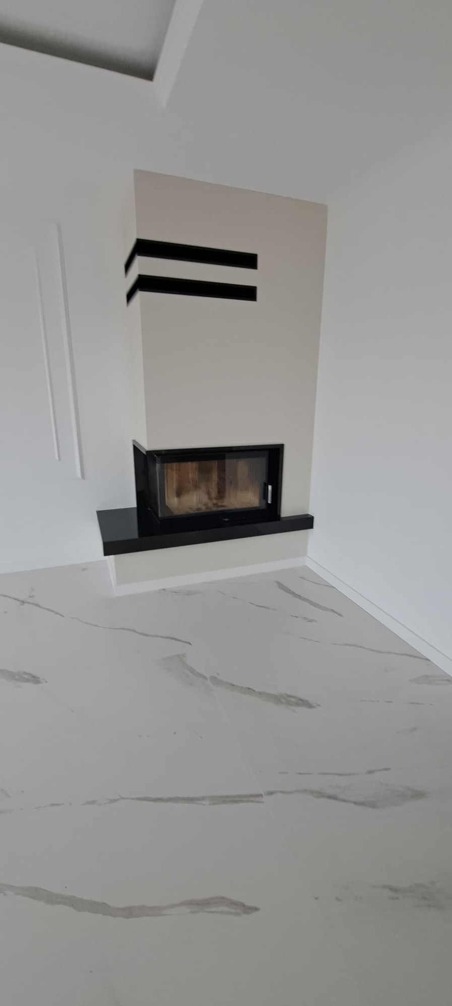 Nowoczesny kominek z czarnymi akcentami w minimalistycznym wnętrzu z marmurową podłogą. Widoczne detale wykończenia i geometryczne zdobienia.