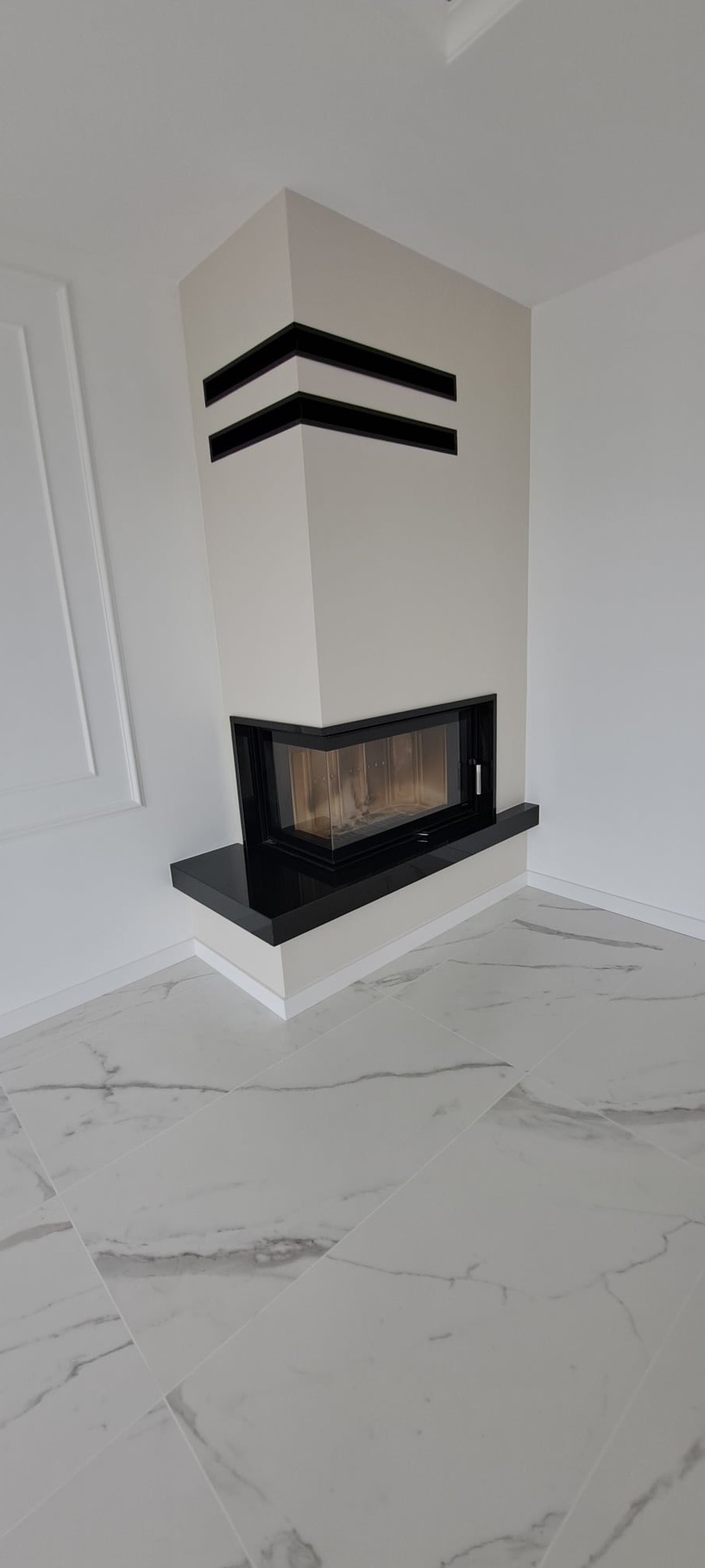 Nowoczesny kominek narożny z czarnymi akcentami, wykończony białymi płytkami podłogowymi z marmurowym wzorem. Minimalistyczny design.