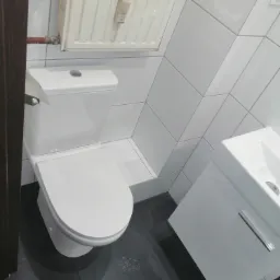 Wykończona łazienka z białymi płytkami ułożonymi pod kątem, szarą podłogą, białą toaletą i umywalką z szafką; widoczny grzejnik z regulatorem temperatury.