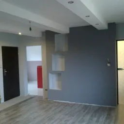 Wnętrze w trakcie wykończenia: szara ściana z wnękami, częściowo ułożona podłoga z paneli imitujących drewno, czarne drzwi po lewej stronie i drzwi z matowego szkła po prawej, widoczne schody w tle.