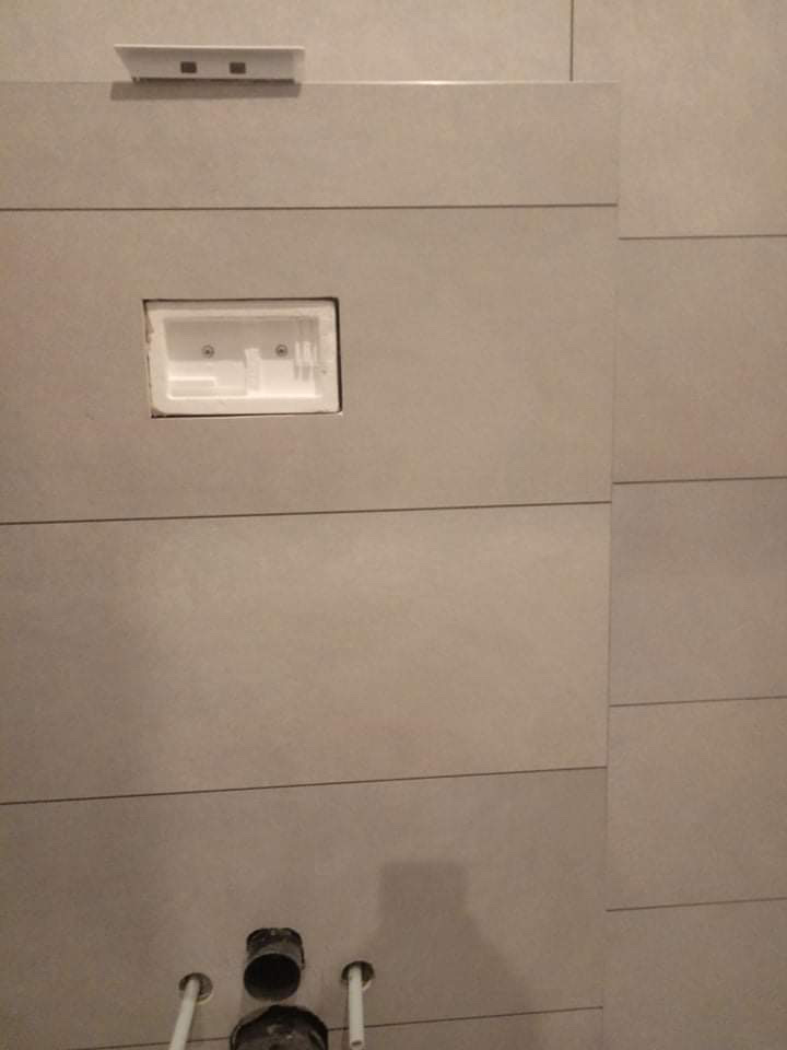 Szare płytki ścienne w łazience, z otworem na stelaż podtynkowy WC i widocznymi rurami instalacyjnymi w dolnej części.