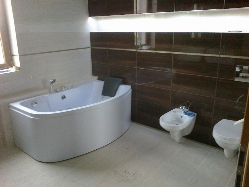 Wykończona łazienka z białą wanną narożną z hydromasażem, bidetem i toaletą, brązowe płytki na ścianie, beżowe płytki na podłodze i ścianie przy oknie.