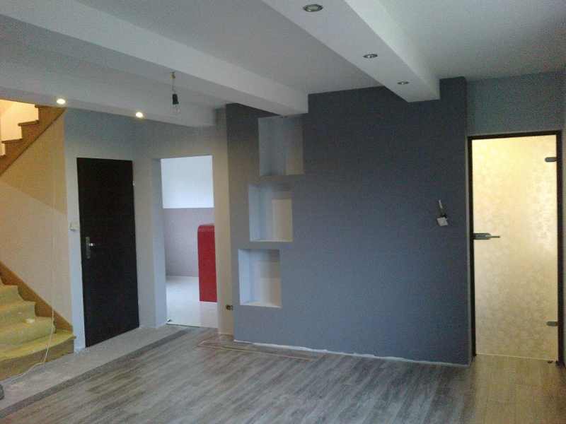 Wnętrze w trakcie wykończenia: szara ściana z wnękami, częściowo ułożona podłoga z paneli imitujących drewno, czarne drzwi po lewej stronie i drzwi z matowego szkła po prawej, widoczne schody w tle.