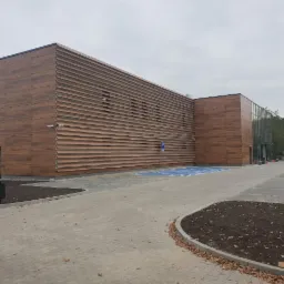 Nowoczesny budynek z elewacją z paneli imitujących drewno, z poziomymi przesłonami na części ściany, widoczne miejsce parkingowe dla niepełnosprawnych i fragment placu z kostki brukowej