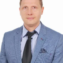 Portret mężczyzny w błękitnej marynarce, czarnym krawacie i wzorzystą poszetką. Uśmiechnięty, na białym tle.