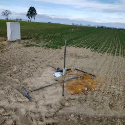 Zestaw do sondowania gruntu na polu uprawnym z widocznym białym kontenerem w tle i notatnikiem obok urządzenia.