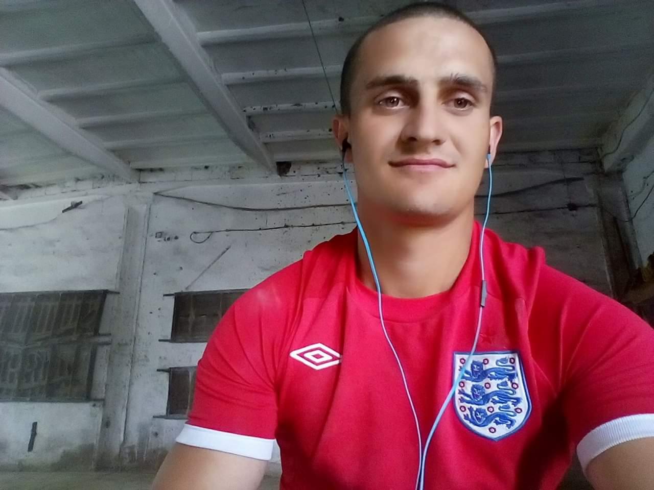 Mężczyzna w czerwonej koszulce z godłem Anglii i słuchawkami na tle białego, surowego sufitu i ściany.