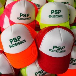 NITKA DAWID KOMARNICKI - Stos czapek z daszkiem typu trucker z białym przodem i kolorową siatką (neonowy żółty, pomarańczowy, czerwony, różowy) z zielonym nadrukiem 'PSP Gołuszowice'.