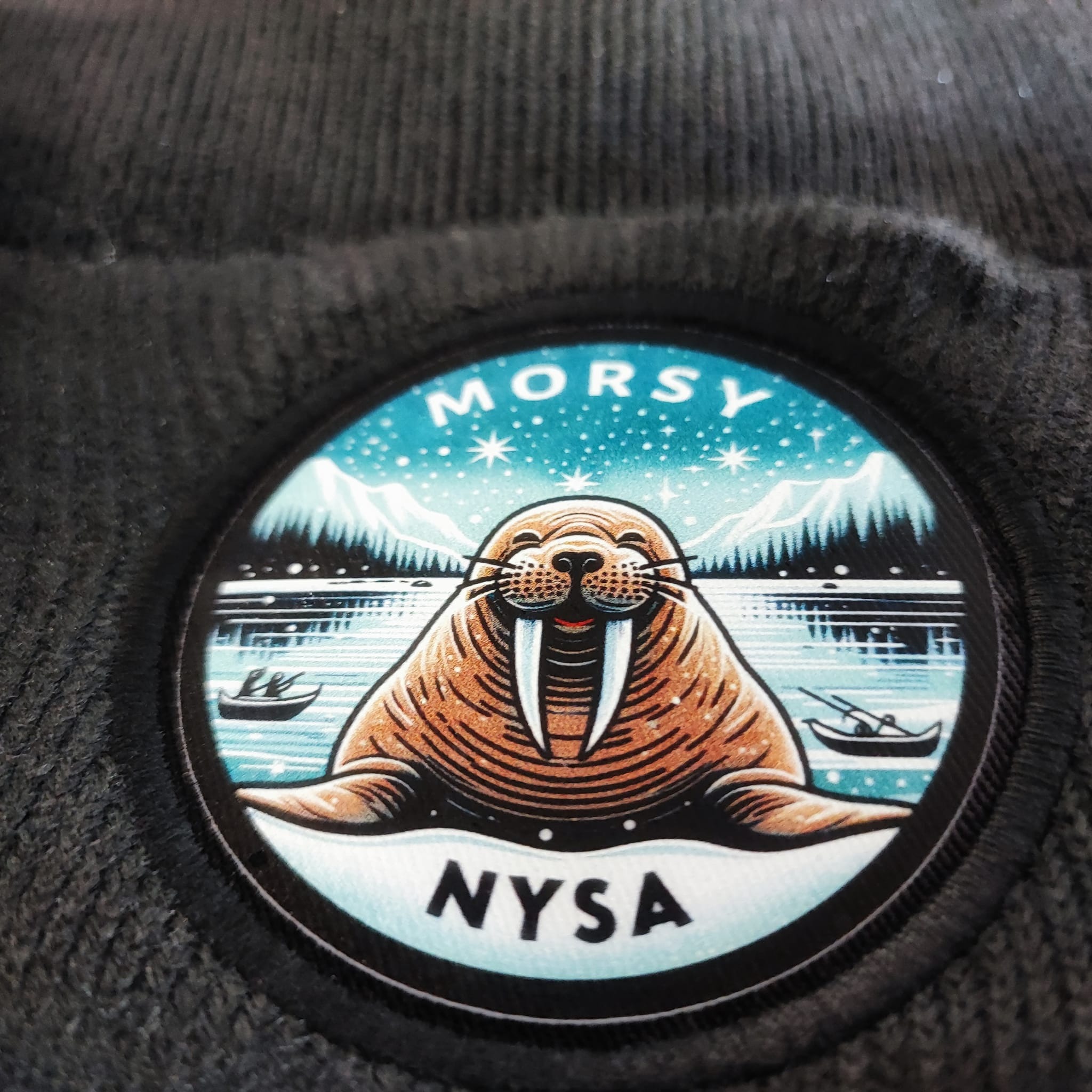 Czarny materiał z naszywką przedstawiającą morsa na tle zimowego krajobrazu z górami, śniegiem i łódkami, z napisem 'MORSY NYSA'.