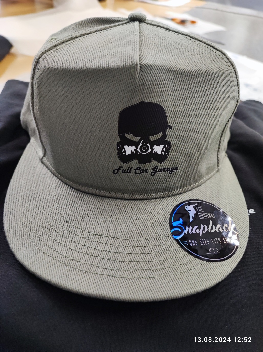 Szara czapka typu snapback z czarnym nadrukiem czaszki w masce gazowej i napisem 'Full Car Garage', obok naklejka 'The Original Snapback'.