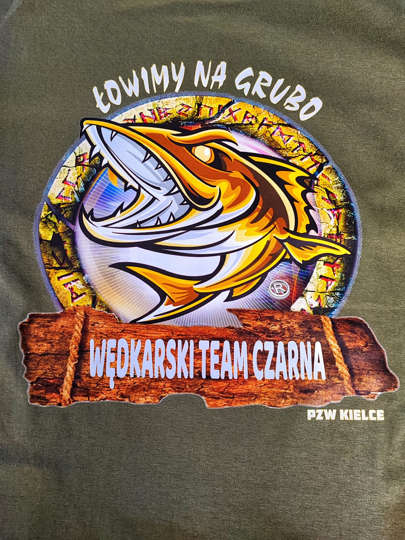 Zielona koszulka z nadrukiem przedstawiającym stylizowaną rybę z otwartą paszczą i napisem 'Łowimy na grubo', poniżej napis 'Wędkarski Team Czarna' na tle imitującym drewnianą tabliczkę, oraz 'PZW...