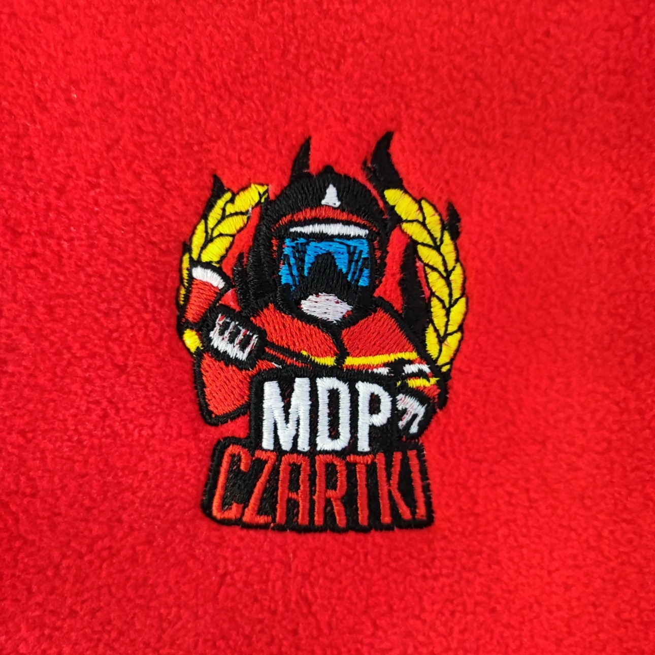Czerwona tkanina frotte z wyhaftowanym kolorowym logo MDP Czartki przedstawiającym postać w hełmie strażackim otoczoną wieńcem laurowym.