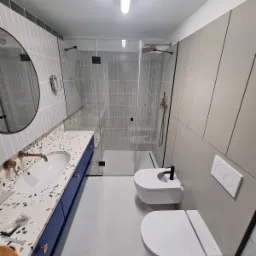 Nowoczesna łazienka z kabiną prysznicową typu walk-in, umywalką nablatową na szafce w kolorze granatowym z blatem lastryko, wiszącą miską WC i bidetem. Armatura w kolorze miedzianym. Na blacie...