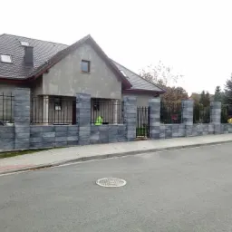 Nowoczesne ogrodzenie z szarej kostki modułowej z metalowymi przęsłami, oddzielające posesję z domem w stanie surowym od ulicy, z widocznym włazem kanalizacyjnym na jezdni.