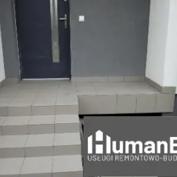 Wyłożone jasnymi płytkami schody wejściowe do domu z ciemnymi drzwiami, szara ściana, logo firmy budowlanej w dolnej części zdjęcia.