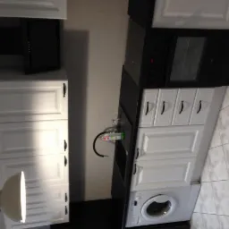 Biała zabudowa kuchenna z czarnym blatem, piekarnikiem i okapem, z pralką w zabudowie pod blatem, widok na szafki górne i dolne z ciemnymi uchwytami.