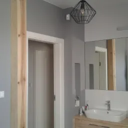 Nowoczesna łazienka w odcieniach szarości z drewnianym akcentem kolumny, lustrem nad umywalką odbijającym fragment ceglanej ściany i grzejnik z ręcznikami, oraz geometryczną lampą wiszącą.