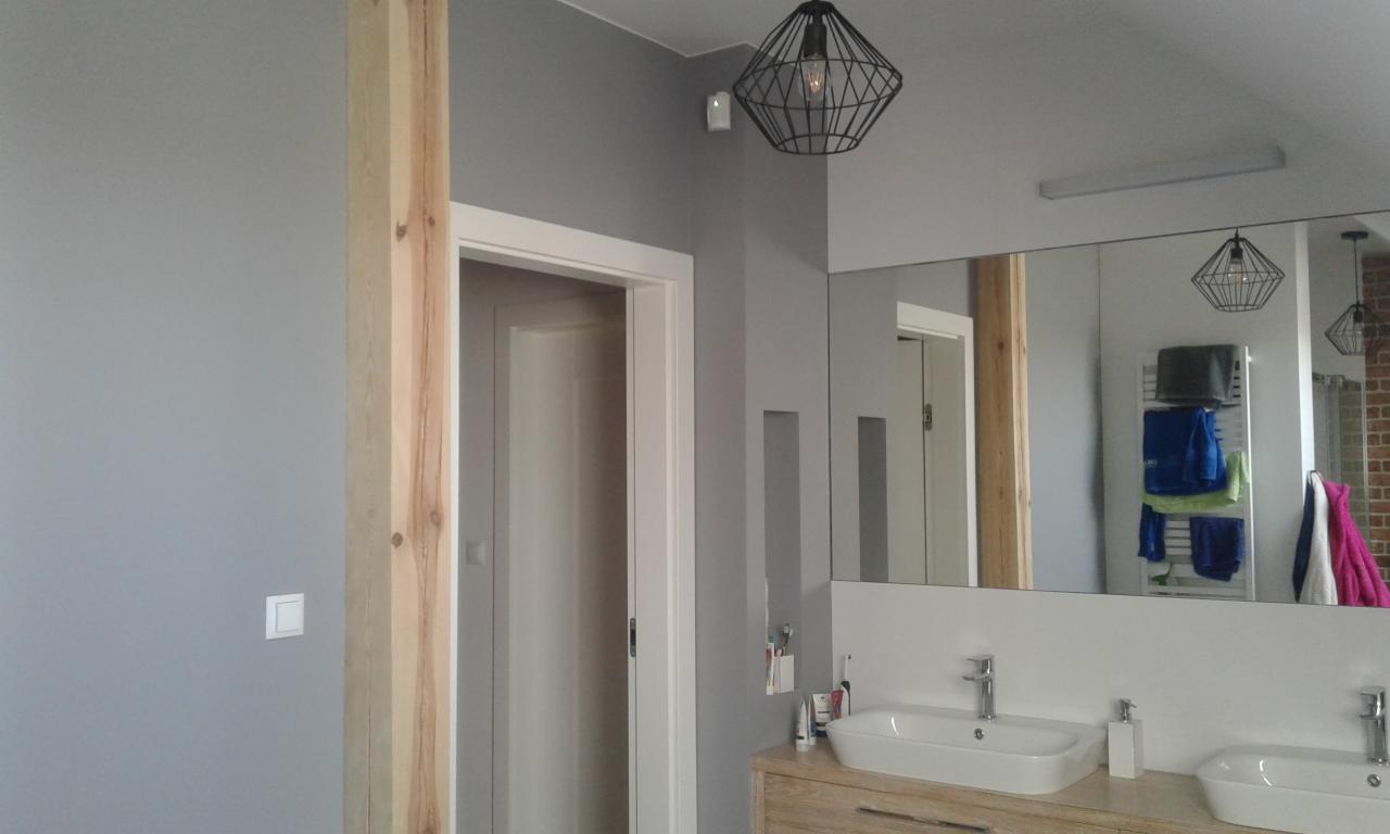 Nowoczesna łazienka w odcieniach szarości z drewnianym akcentem kolumny, lustrem nad umywalką odbijającym fragment ceglanej ściany i grzejnik z ręcznikami, oraz geometryczną lampą wiszącą.