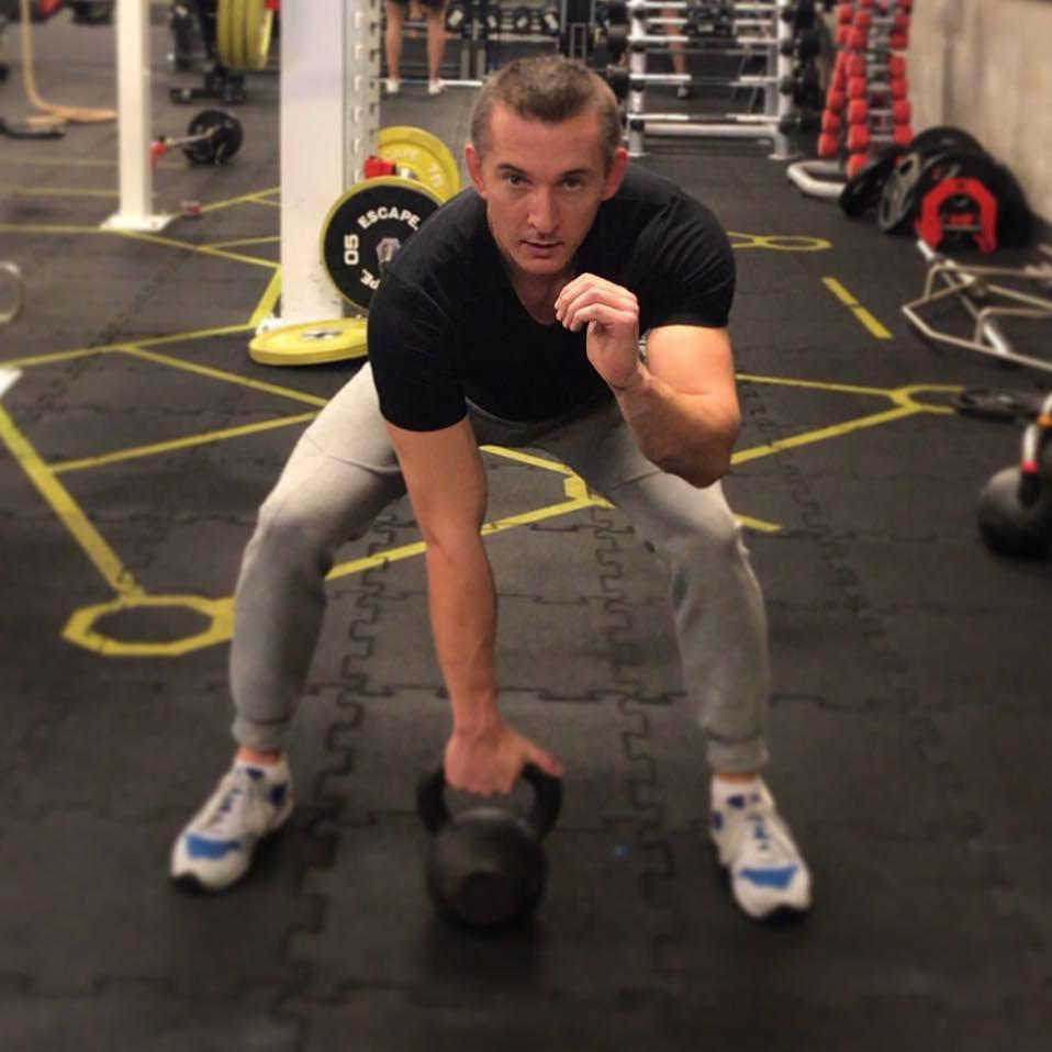 Mężczyzna w szarej bluzie dresowej i czarnej koszulce podnosi kettlebell w siłowni; widoczne obciążenia i sprzęt do ćwiczeń w tle.