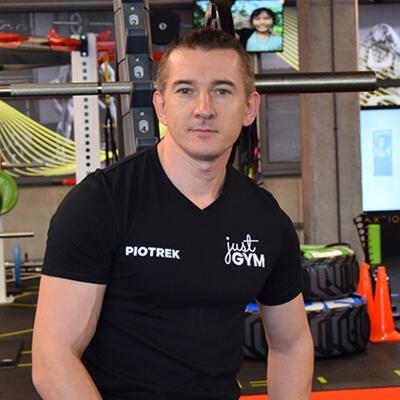 Mężczyzna w czarnej koszulce z napisem 'Piotrek' i logo 'Just Gym' na tle siłowni z maszynami do ćwiczeń i oponami