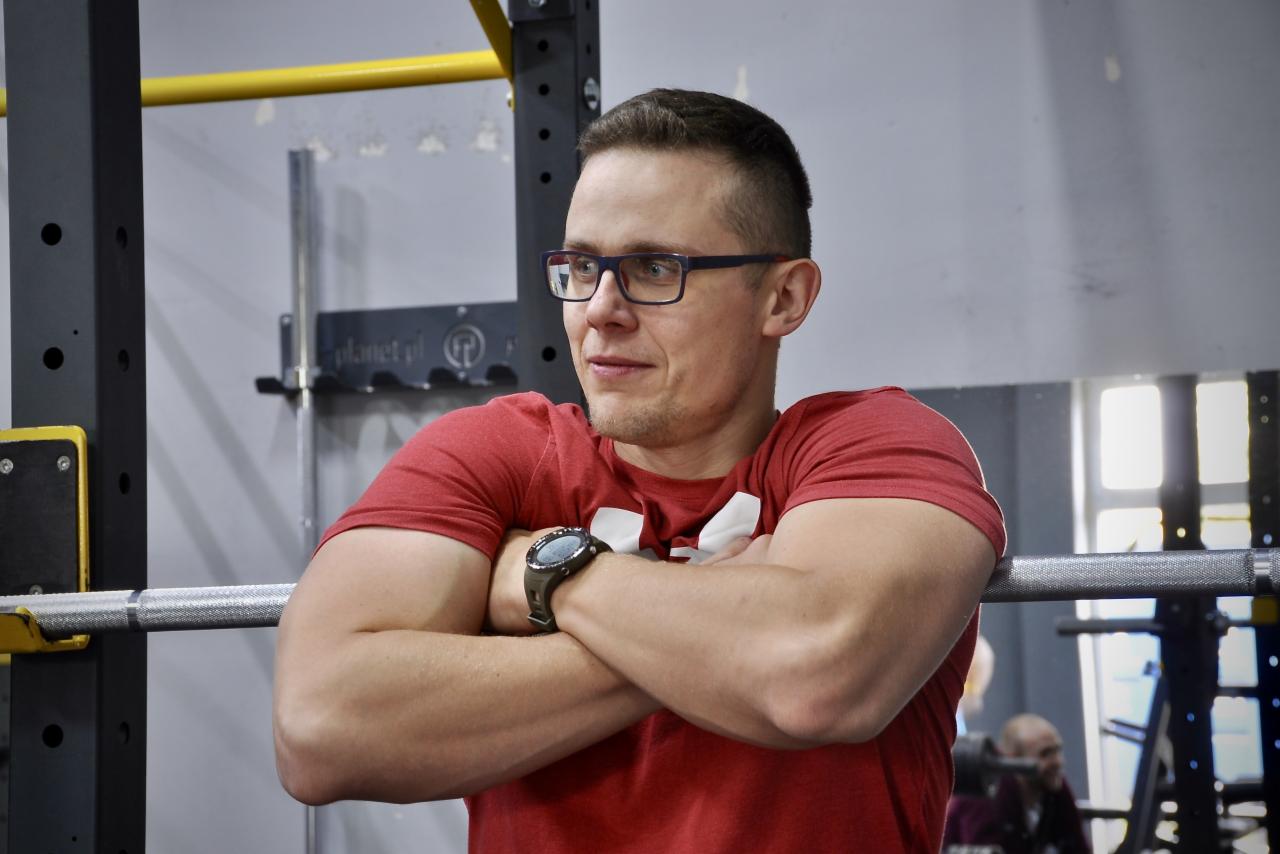 Trener personalny w czerwonej koszulce, z założonymi rękami, w tle sprzęt siłowni. Mężczyzna w okularach, z zegarkiem na ręku, spogląda w bok.