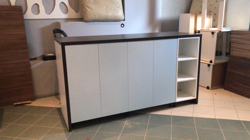 Biała szafka z czarnym blatem i otwartą półką z trzema przegródkami, stojąca na podłodze wyłożonej kwadratowymi płytkami, w tle widoczne elementy meblowe i płyty.