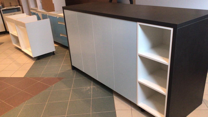 Częściowo zmontowana szafka kuchenna z szarymi frontami i czarnym blatem, widoczne białe półki i podłoga z płytek w geometryczne wzory. W tle inne elementy zabudowy kuchennej.