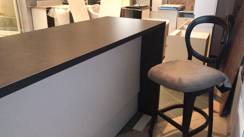 Wysoki blat kuchenny w odcieniach szarości, z czarnym obramowaniem, ustawiony w pomieszczeniu w trakcie montażu, obok stoi krzesło barowe z ciemnego drewna z szarym siedziskiem, w tle widoczne...