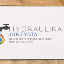 Logo firmy hydraulicznej Jurzysta na białej kartce, otoczone narzędziami: miarka, poziomica, suwmiarka, okulary, kask ochronny i zwinięte plany instalacji na tle drewnianego blatu.