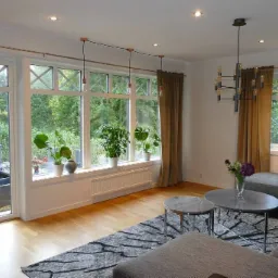 Jasny salon z dużym oknem z białymi ramami, przez które widać zieleń ogrodu, rośliny doniczkowe na parapecie, szare zasłony i nowoczesny żyrandol.