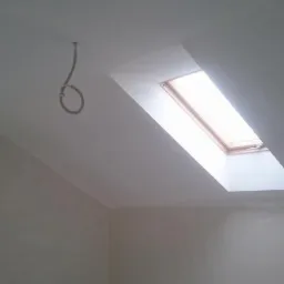 Pokój na poddaszu w trakcie wykańczania, ściany i sufit świeżo pokryte gładzią, widoczne okno dachowe i wystający kabel elektryczny.