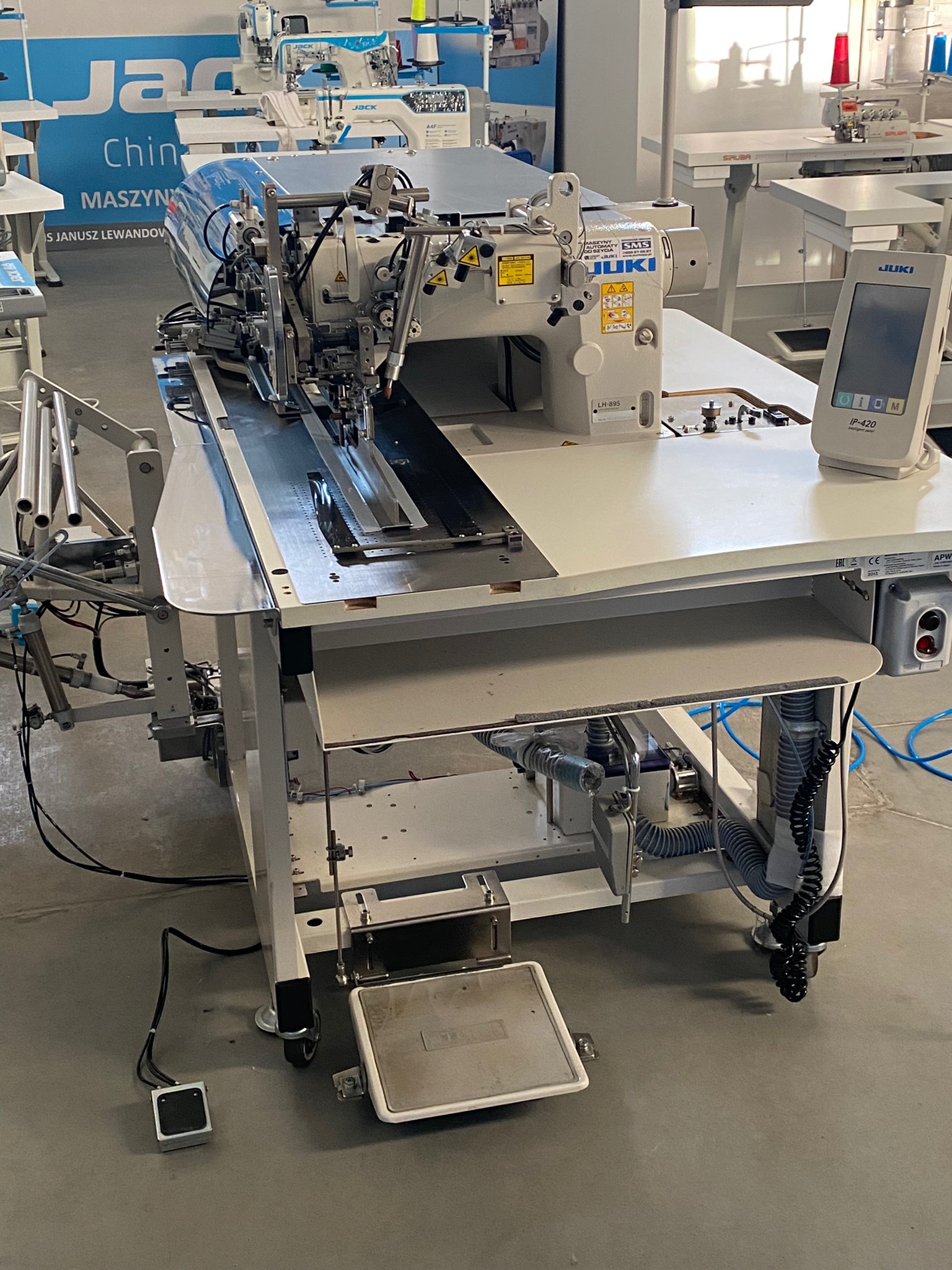 Profesjonalna, przemysłowa maszyna do szycia JUKI LH-895 z panelem sterowania IP-420 na białym stole, widoczny pedał nożny i elementy pneumatyczne pod blatem. W tle inne maszyny szwalnicze marek...