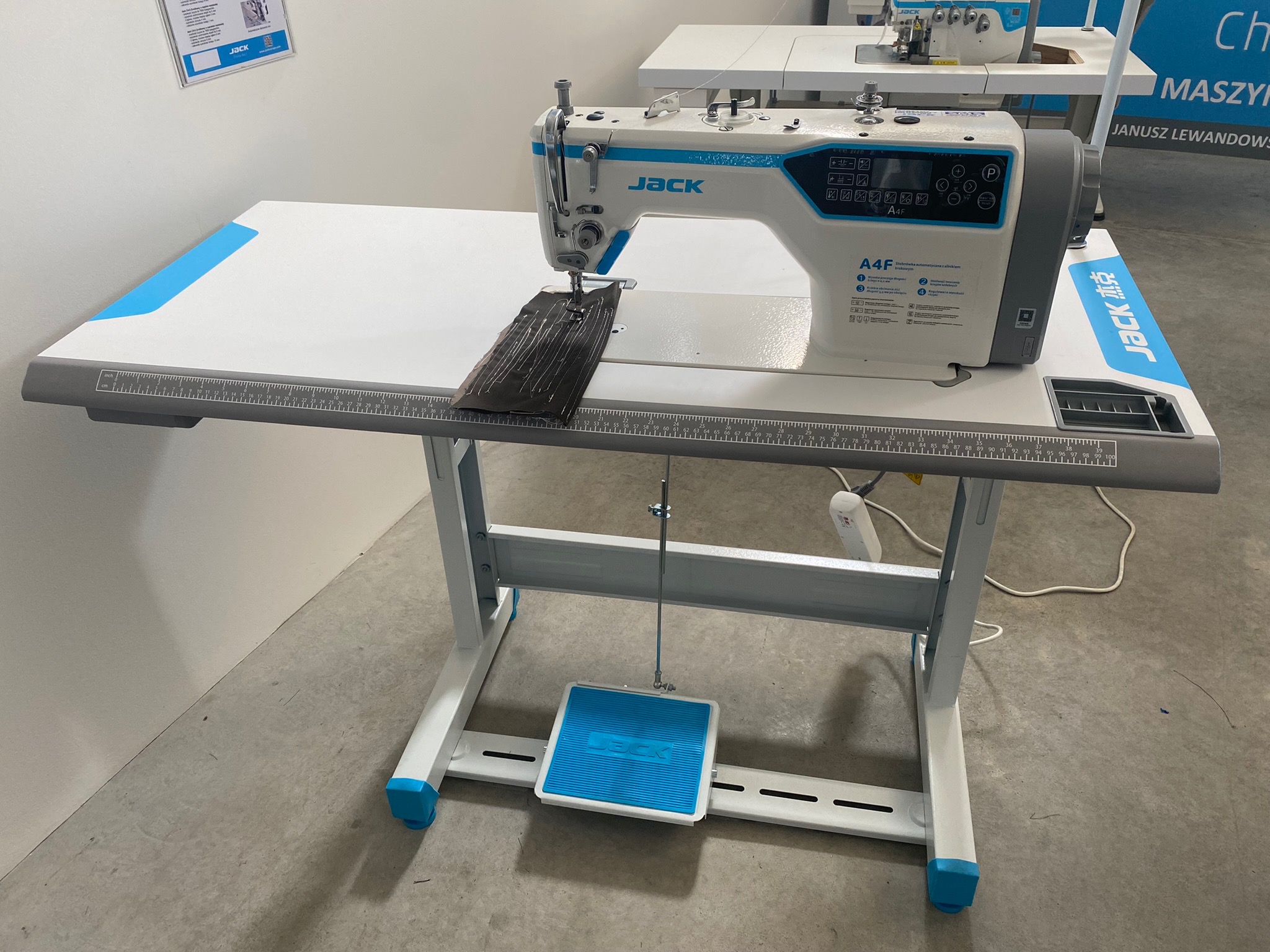 Przemysłowa maszyna do szycia Jack A4F na stole z miarką, szyjąca ciemny materiał w jasnym pomieszczeniu.