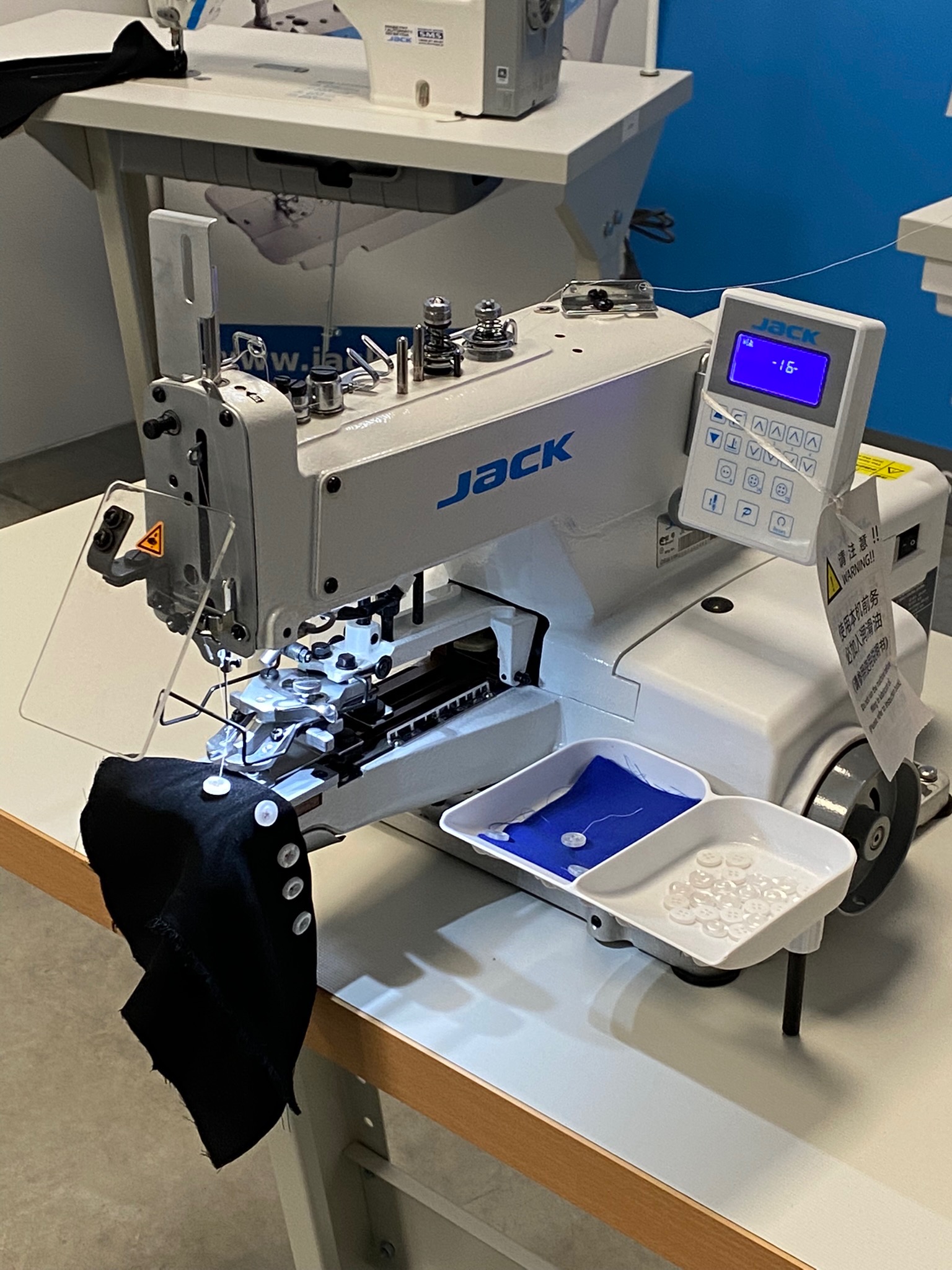 Przemysłowa maszyna do szycia guzików marki Jack, z zamocowanym czarnym materiałem z przyszytymi guzikami i pojemnikiem z zapasem guzików, ustawiona na stole. Widoczny panel sterowania...