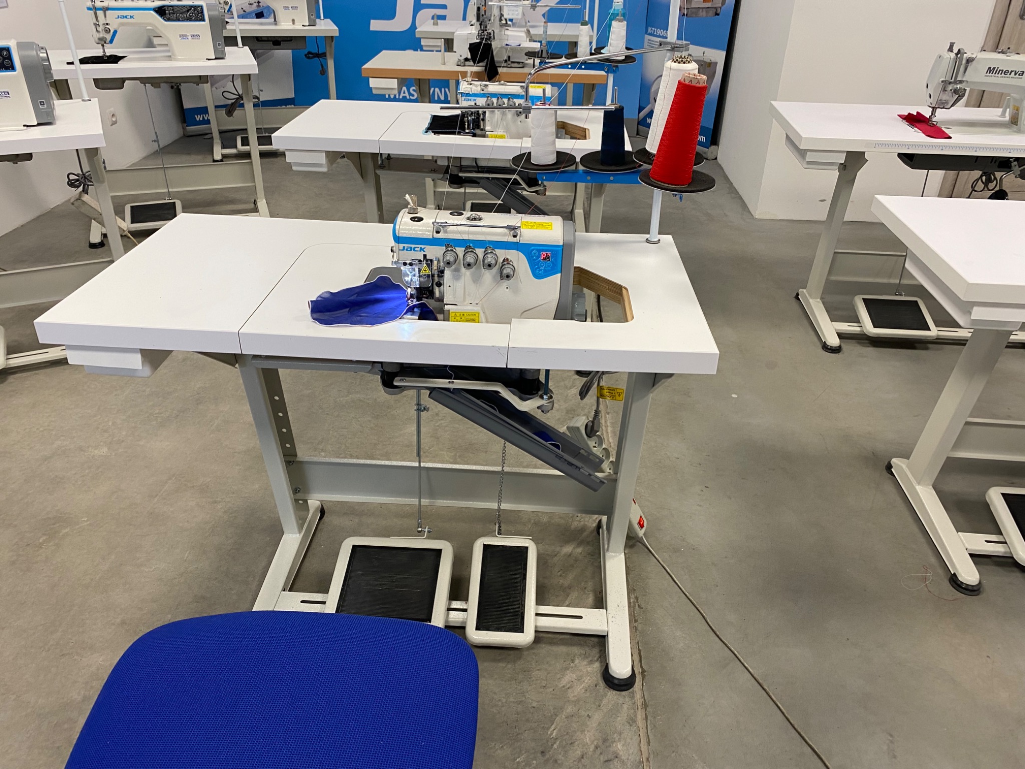 Overlock przemysłowy marki Jack z niebieskim materiałem pod stopką, widoczne szpule nici w kolorze czerwonym, białym i granatowym, stanowisko pracy z pedałami nożnymi i niebieskim siedziskiem...