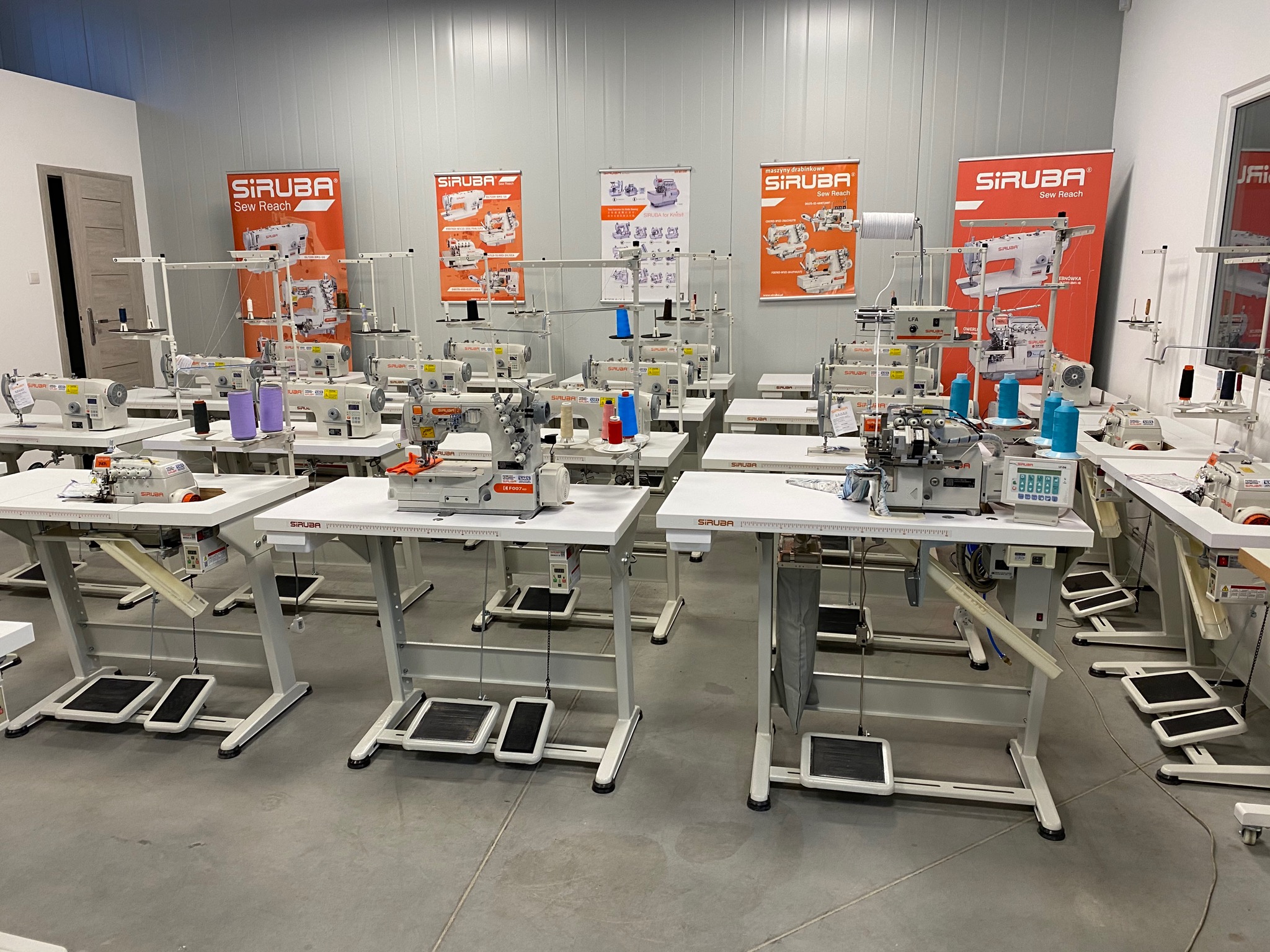 Rząd nowych, białych maszyn szwalniczych marki Siruba, ustawionych na białych stołach z pedałami nożnymi, w jasnym pomieszczeniu z plakatami reklamowymi marki w tle.