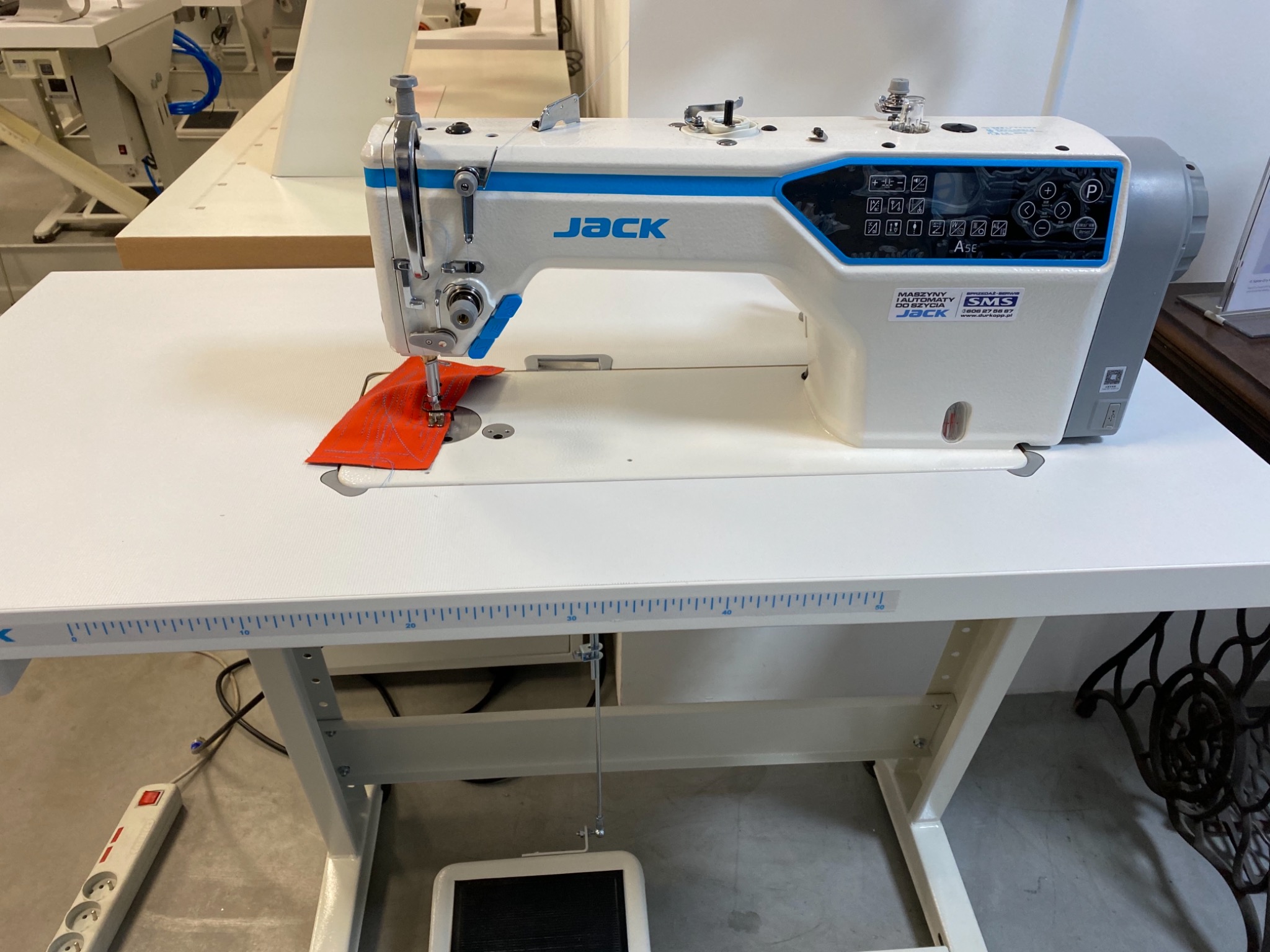 Przemysłowa maszyna do szycia marki Jack, model A5E, z pomarańczowym materiałem pod igłą, widoczny pedał nożny i miarka na blacie stołu.
