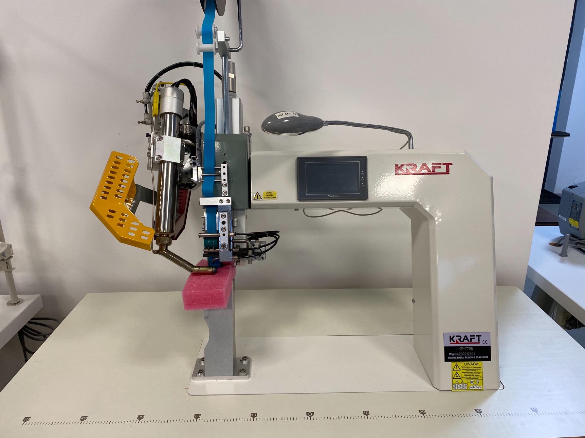 Szczegółowe ujęcie przemysłowej maszyny szwalniczej marki Kraft, model KF-7705, z widocznym wyświetlaczem Samkoon i żółtą osłoną, ustawionej na stole z podziałką centymetrową.
