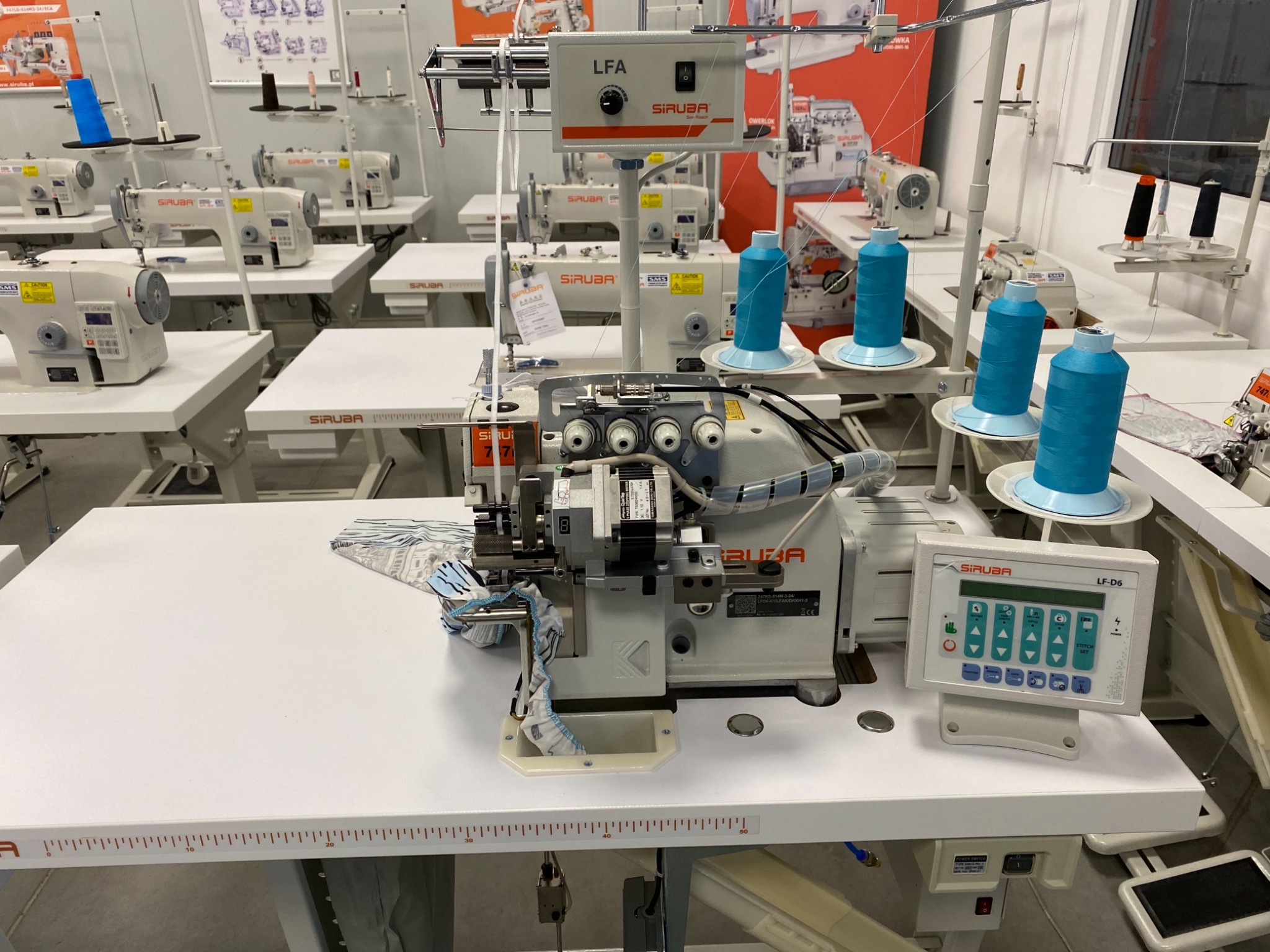 Szwalnia: Profesjonalny overlock przemysłowy marki Siruba LF-D6 z nawleczonymi niebieskimi nićmi, gotowy do szycia, na tle innych maszyn szwalniczych.