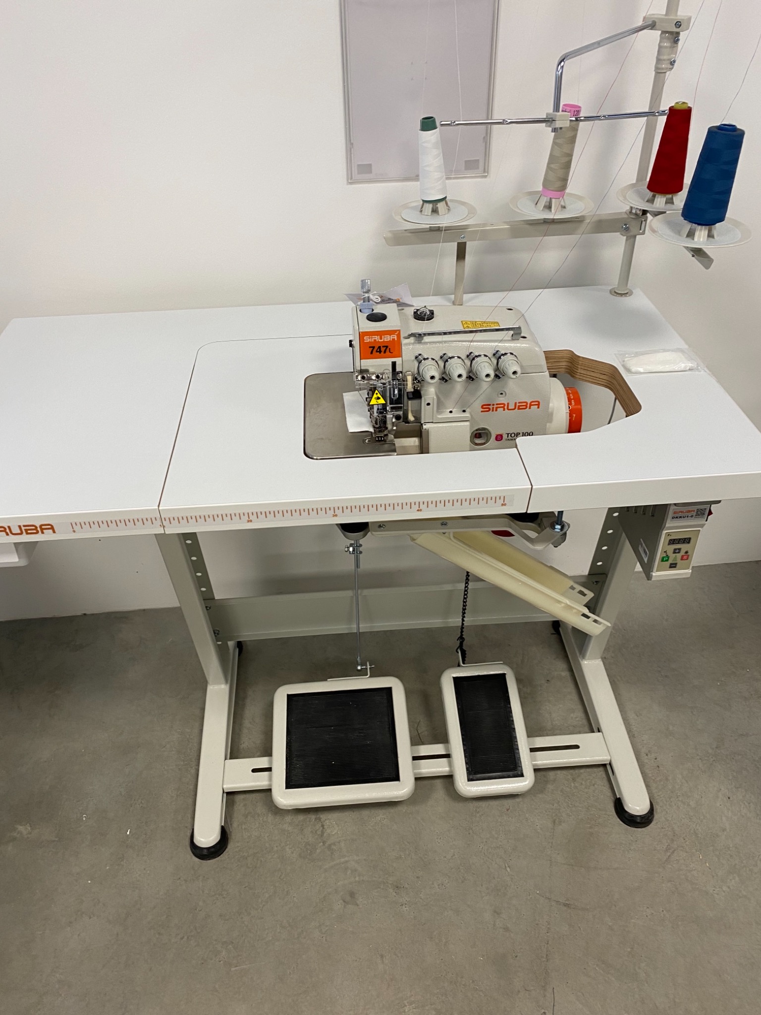 Profesjonalna, biała owerlokowa maszyna do szycia marki Siruba, model TOP100, z czterema nałożonymi szpulkami nici w kolorach białym, beżowym, czerwonym i niebieskim, stojąca na białym stole z miarką.