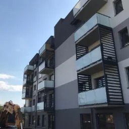 Nowoczesny budynek wielorodzinny z balkonami zabezpieczonymi szklanymi balustradami, widoczne prace budowlane w tle z użyciem koparki.