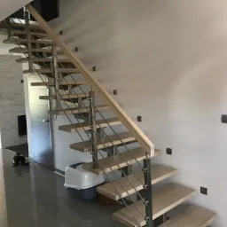 Schody drewniane z metalowymi barierkami, minimalistyczny design, widoczny koci transporter pod schodami, szare płytki podłogowe.