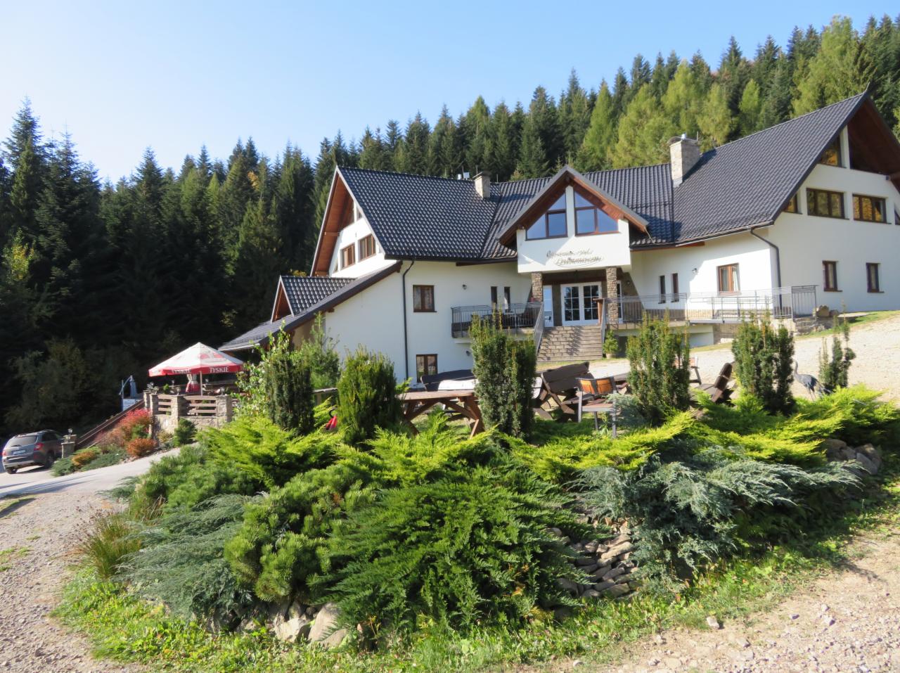 Dwupiętrowy budynek restauracji z ciemnym dachem, otoczony zielenią i drzewami, widoczny parasol Tyskie.