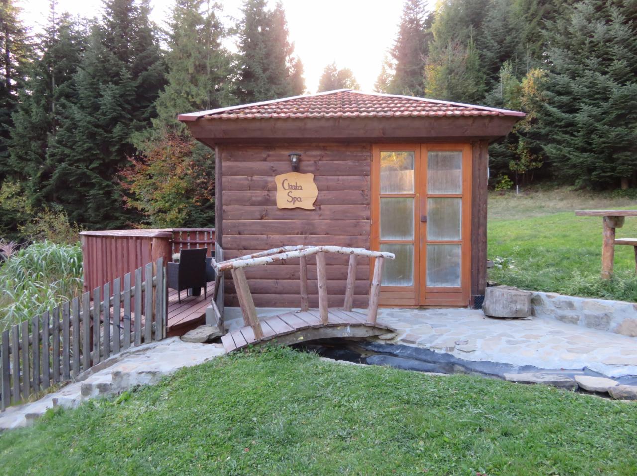 Drewniana chata spa z mostkiem z białej brzozy nad strumieniem, otoczona zielenią i drzewami iglastymi.